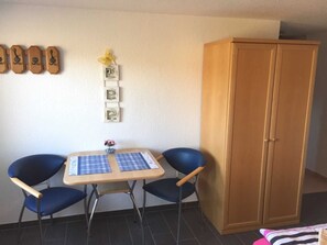 Une petite pièce avec une armoire en bois, deux chaises bleues, une table recouverte d’une nappe à carreaux et un mur orné de tableaux et d’une fleur jaune.