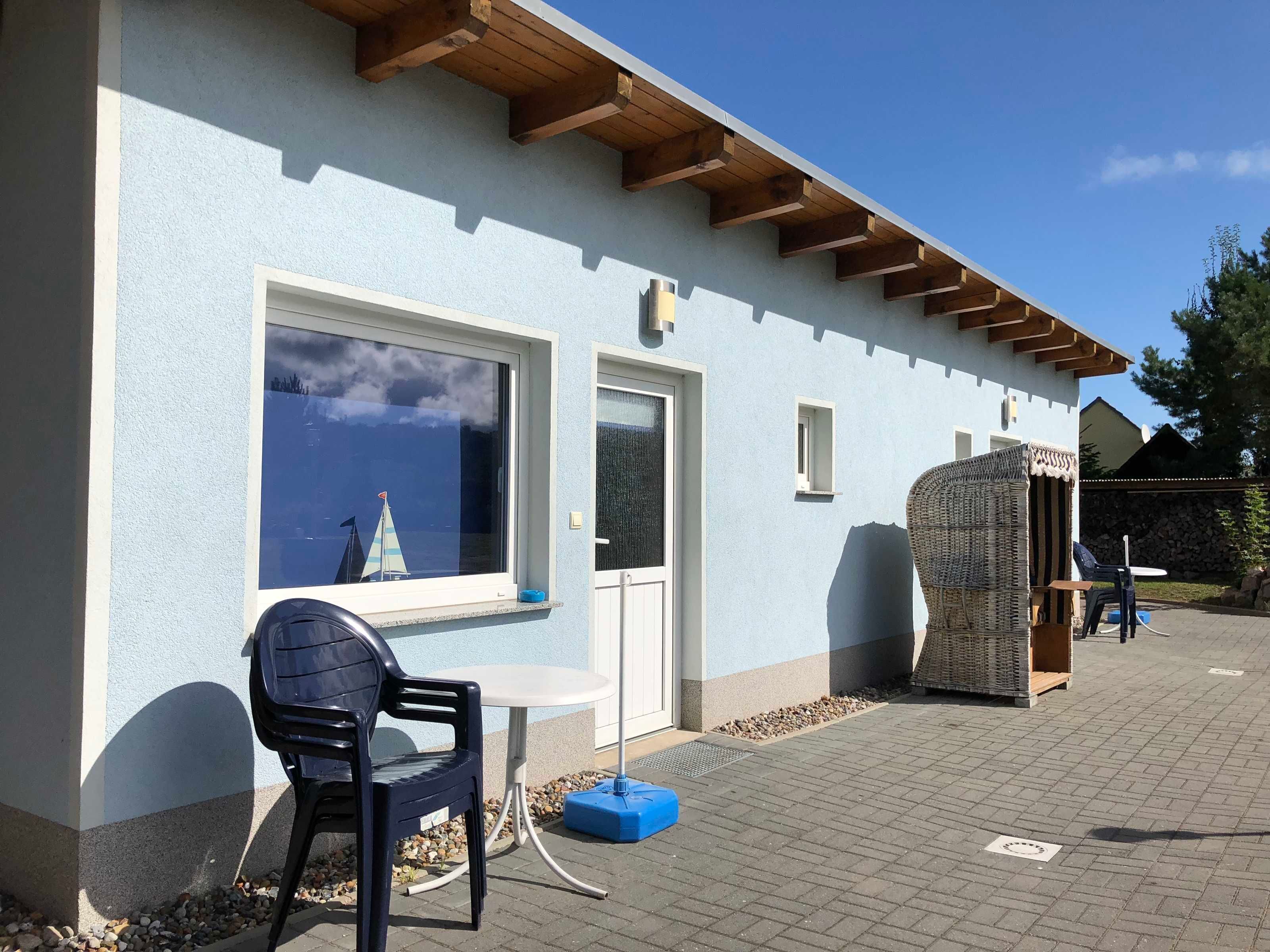 Ein Gebäude mit hellblauem Anstrich, Holzbalken am Dach, einem Fenster mit einem Segelmodell, einer Tür und einem kleinen Tisch mit Stuhl davor.