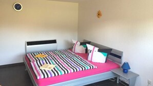 Un dormitorio con cama, mesita de noche con lámpara, un reloj de pared y una pequeña decoración floral.
