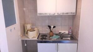 Una pequeña cocina americana con tostadora, un lavamanos y una estufa. Hay un cubo de basura y un armario sobre el lavamanos.