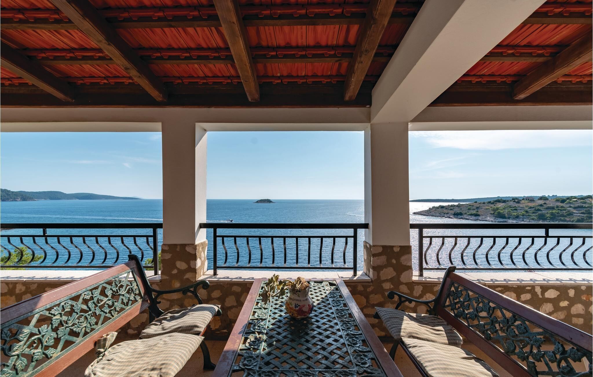 A balcony with two wooden benches and a table, overlooking a coastal view with a distant island.