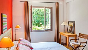 Quarto com cama, luminária de cabeceira, cadeira, mesa pequena com abajur, espelho e janela com cortinas.