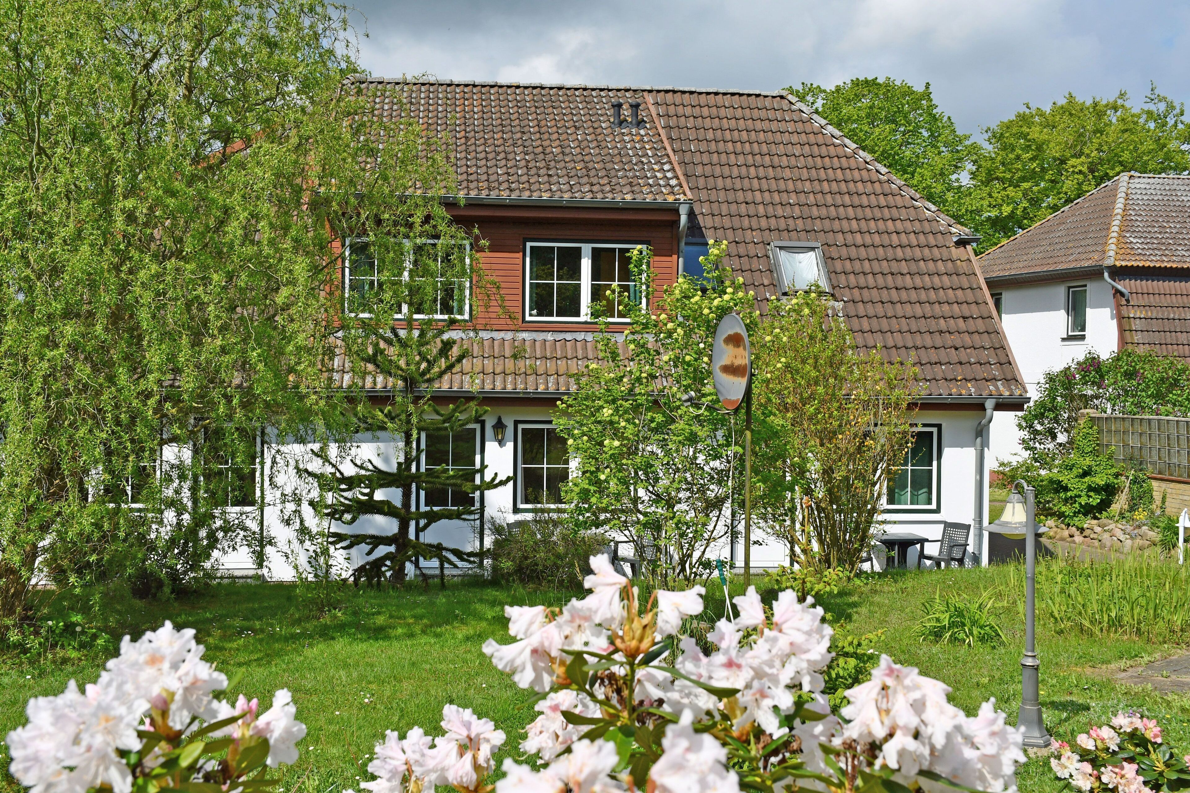 Ein zweistöckiges Haus mit einem Ziegeldach, umgeben von Grünflächen und blühenden Pflanzen.