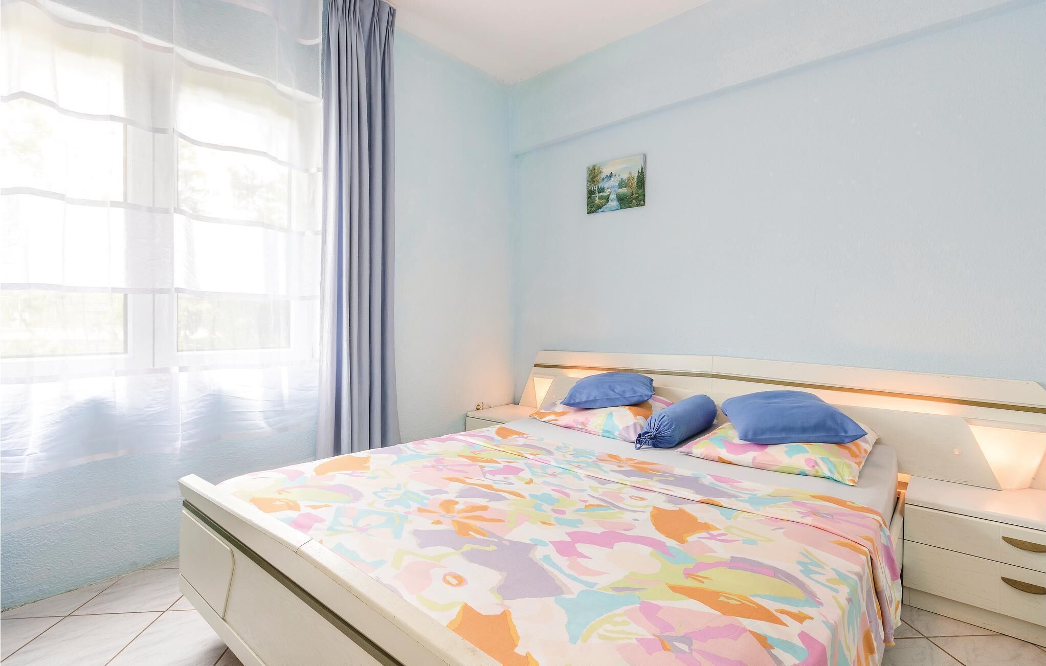 A bedroom with a bed, two bedside tables, a nightstand, a window with curtains, and a painting on the wall.