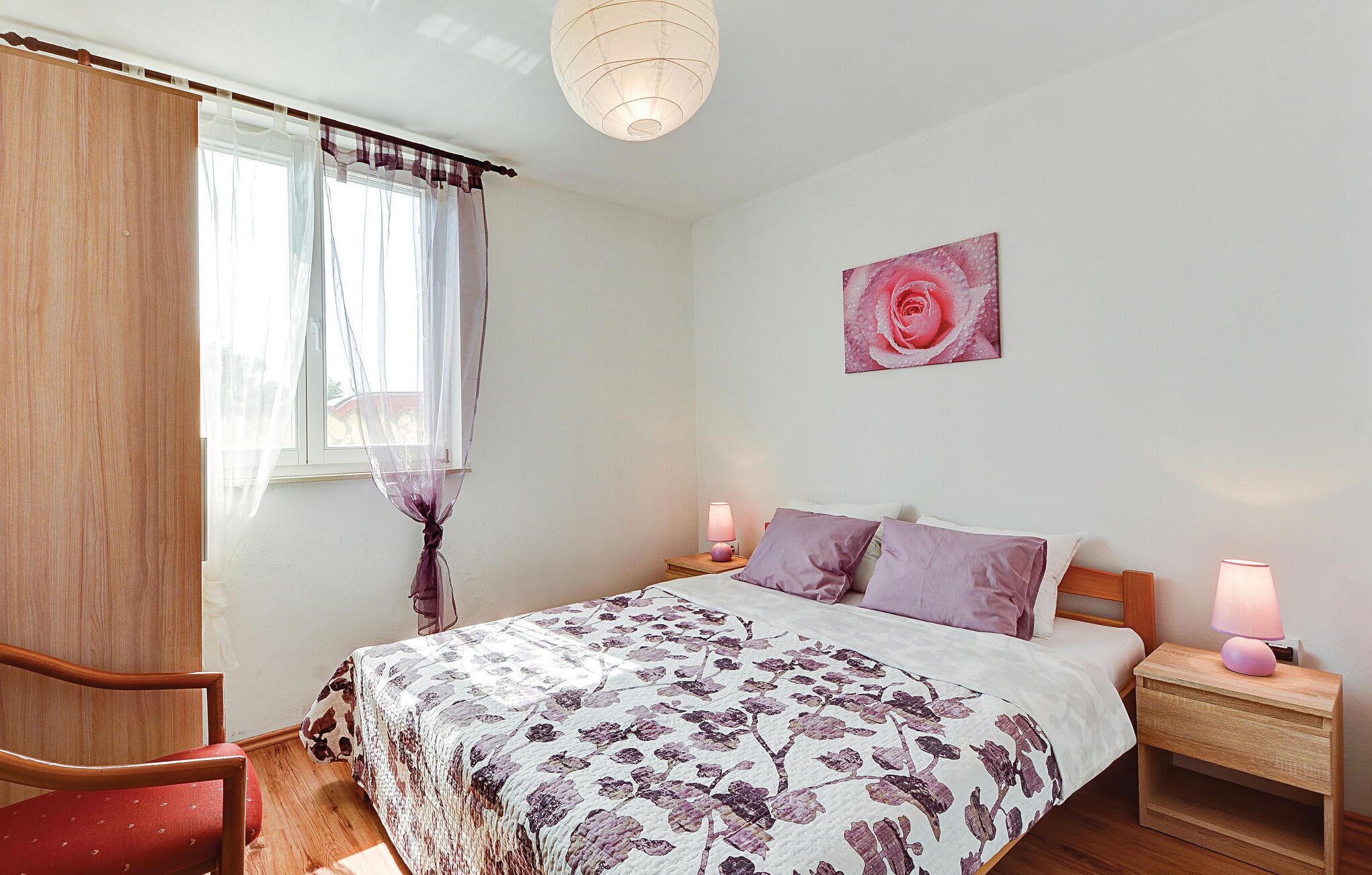Une chambre à coucher avec un lit, deux tables de chevet avec des lampes, une chaise, une fenêtre avec des rideaux légers et un tableau représentant une rose accroché au mur.