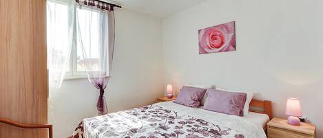 Une chambre à coucher avec un lit, deux tables de chevet avec des lampes, une chaise, une fenêtre avec des rideaux légers et un tableau représentant une rose accroché au mur.