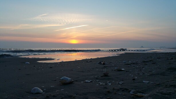 Une plage au coucher du soleil, avec le soleil bas à l’horizon, les vagues qui viennent doucement lécher le rivage, et une jetée qui s’avance dans l’eau.
