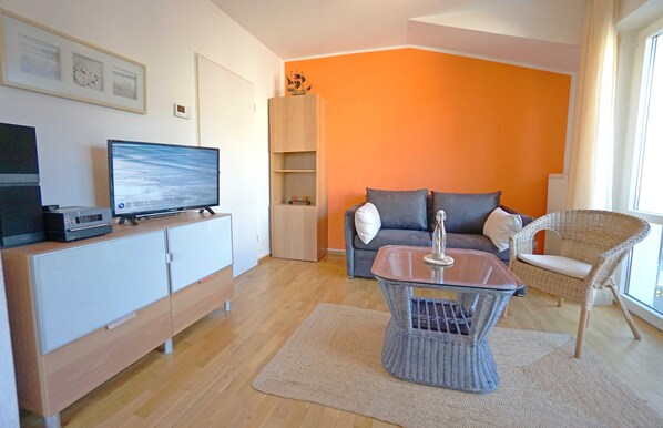 A modern living room with a flat-screen TV, a sofa, a wicker chair, and a glass coffee table.