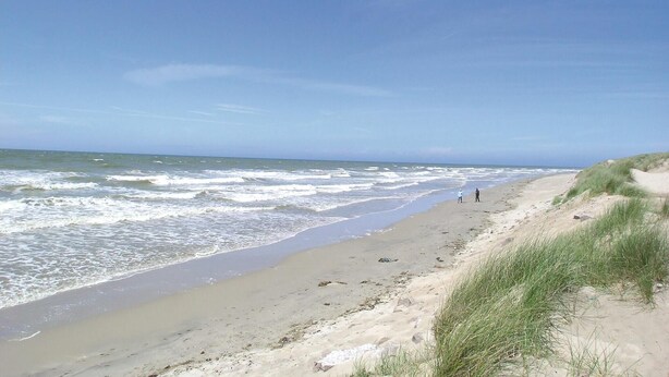Ein Strand mit Wellen, blauem Himmel und zwei Personen, die spazieren gehen.