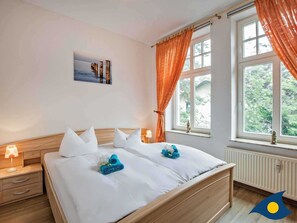 Un dormitorio con cama de madera, ropa de cama blanca, una mesita de noche con lámpara, una ventana con cortinas naranjas y vistas al exterior con vegetación.