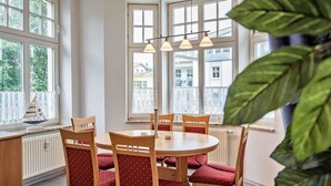 A dining area with a round table and chairs, large windows, and a view of buildings outside. - Villa Malve Whg. 05 - VMa 05 (Heringsdorf)