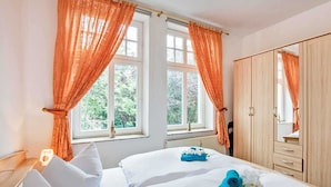 A hotel room with two beds, orange curtains, a wooden wardrobe, and a window with a view of greenery. - Villa Malve Whg. 05 - VMa 05 (Heringsdorf)