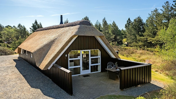 En hytte med stråtækt tag, træfacade, stor glasdør og et lille udendørs siddeområde.