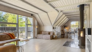 En rummelig loftrum med højt til loftet, en stor skydedør der fører ud til en træterrasse, et hyggeligt opholdsområde med sofa og stole, et spisebord og en brændeovn.