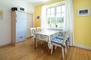 Ein Zimmer mit Holzboden, einem weißen Tisch mit Blumenmuster, vier weißen Stühlen mit grauen Kissen, einer weißen Kommode mit Mikrowelle und einem Fenster mit Blick auf Grünflächen.