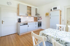 Une cuisine compacte avec des meubles blancs, une cuisinière, un four et un four à micro-ondes. On y trouve une table à manger avec des chaises et une horloge au mur.