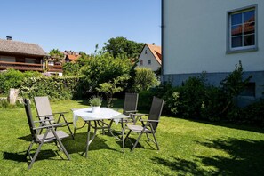 Une terrasse avec une table et des chaises, entourée d’une pelouse bien entretenue et d’un bâtiment à la façade blanche.