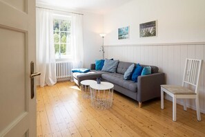Ein Wohnzimmer mit einer Couch, zwei Beistelltischen, einem Sessel, einem Heizkörper, einem Fenster mit Vorhängen und zwei Wandbildern.