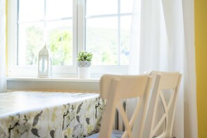 Une table recouverte d’une nappe à motifs, une chaise blanche, une lanterne blanche et une plante en pot posée sur un rebord de fenêtre.