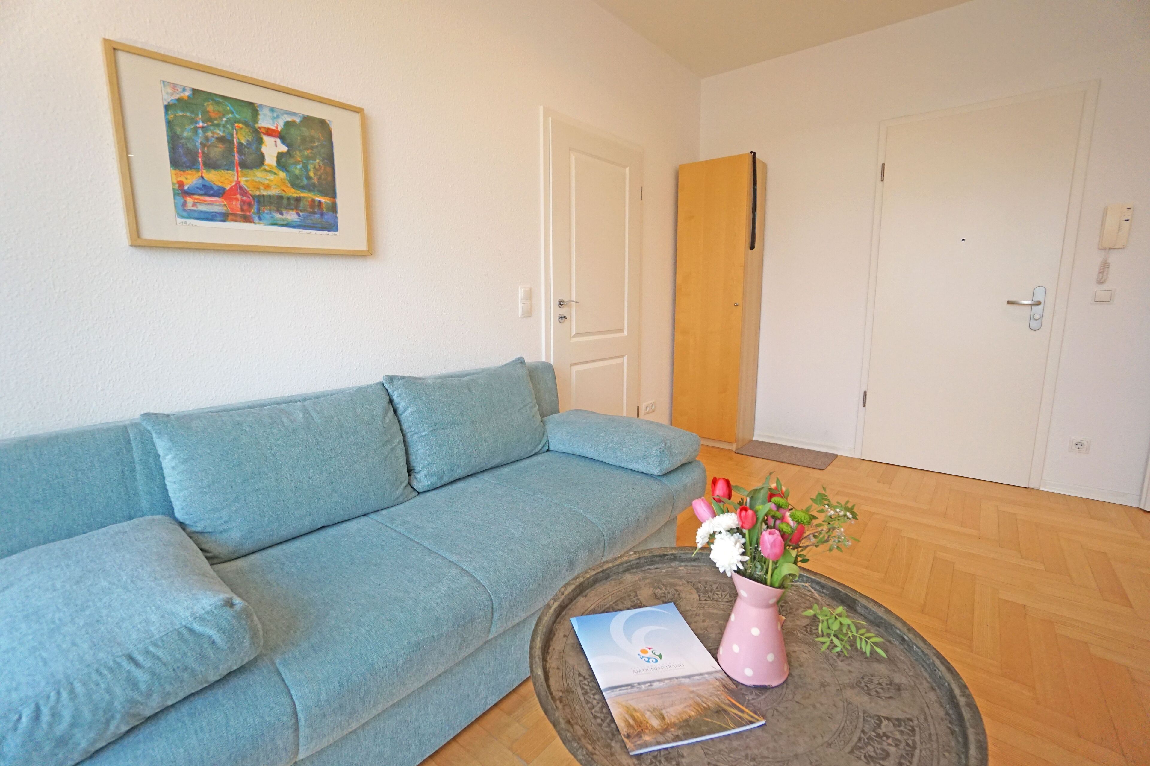 A room with a light blue sofa, a wooden coffee table with a book and a pink vase with flowers, and a framed painting on the wall.