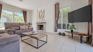 Un salon comprenant un canapé, une table basse, une cheminée, un téléviseur et de grandes fenêtres avec des rideaux.