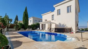 Une maison moderne à deux étages, dotée d’une piscine, entourée de verdure et surplombant un ciel d’un bleu limpide.