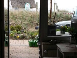 Un chemin pavé de briques mène à un bâtiment dont le toit est recouvert de chaume. Une voiture est garée à proximité, et des conteneurs verts sont posés sur le sol.