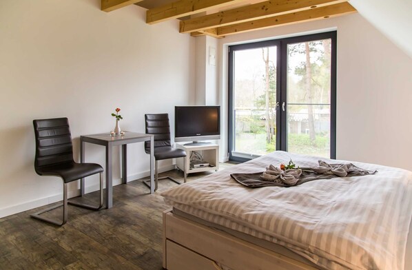 A modern bedroom with a bed, two chairs, a small table, a TV, and a window with a view of greenery.