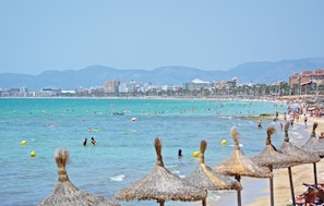 Ein Strand mit Reetdachschirmen, klarem türkisblauem Wasser und einer Stadtlandschaft im Hintergrund.