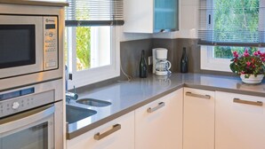A modern kitchen with a built-in microwave, oven, and coffee maker. There is a window with blinds and a potted plant on the counter. - Cozy home in Lluchmajor (Lluchmajor)