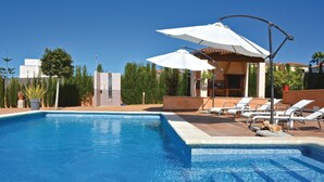 A swimming pool with lounge chairs and umbrellas, surrounded by greenery and a tiled deck. - Cozy home in Lluchmajor (Lluchmajor)