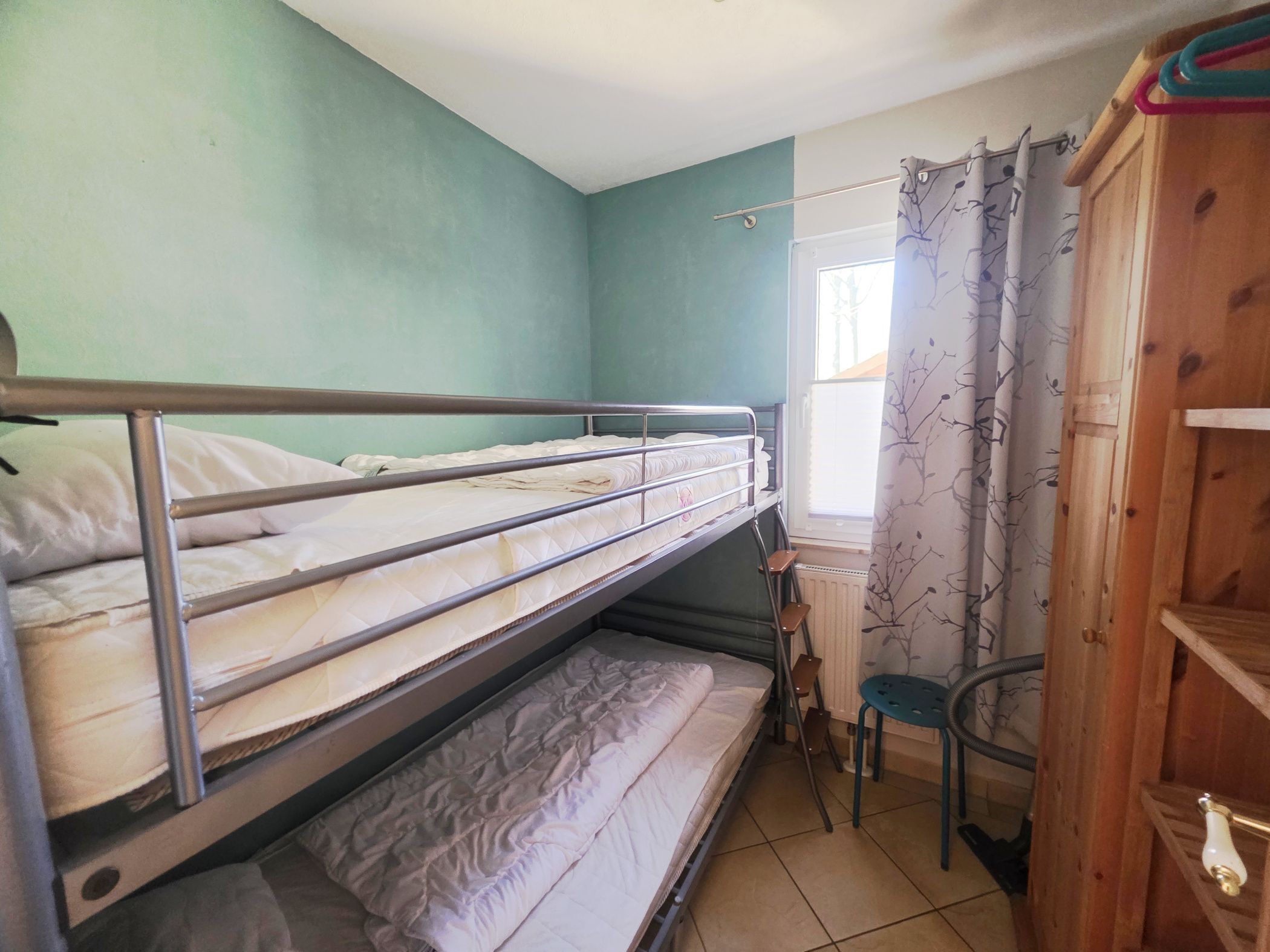 A bunk bed room with a window, a radiator, and a wooden wardrobe.