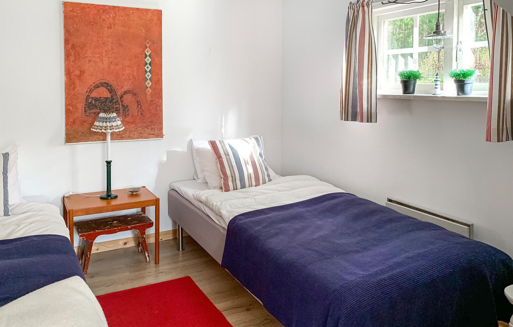 Une chambre avec deux lits simples, une table de chevet, une lampe, un tapis rouge et une fenêtre avec des rideaux.