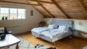 A bedroom with a sloped wooden ceiling, a bed with white bedding, a chair, a small table, and a window with plants.