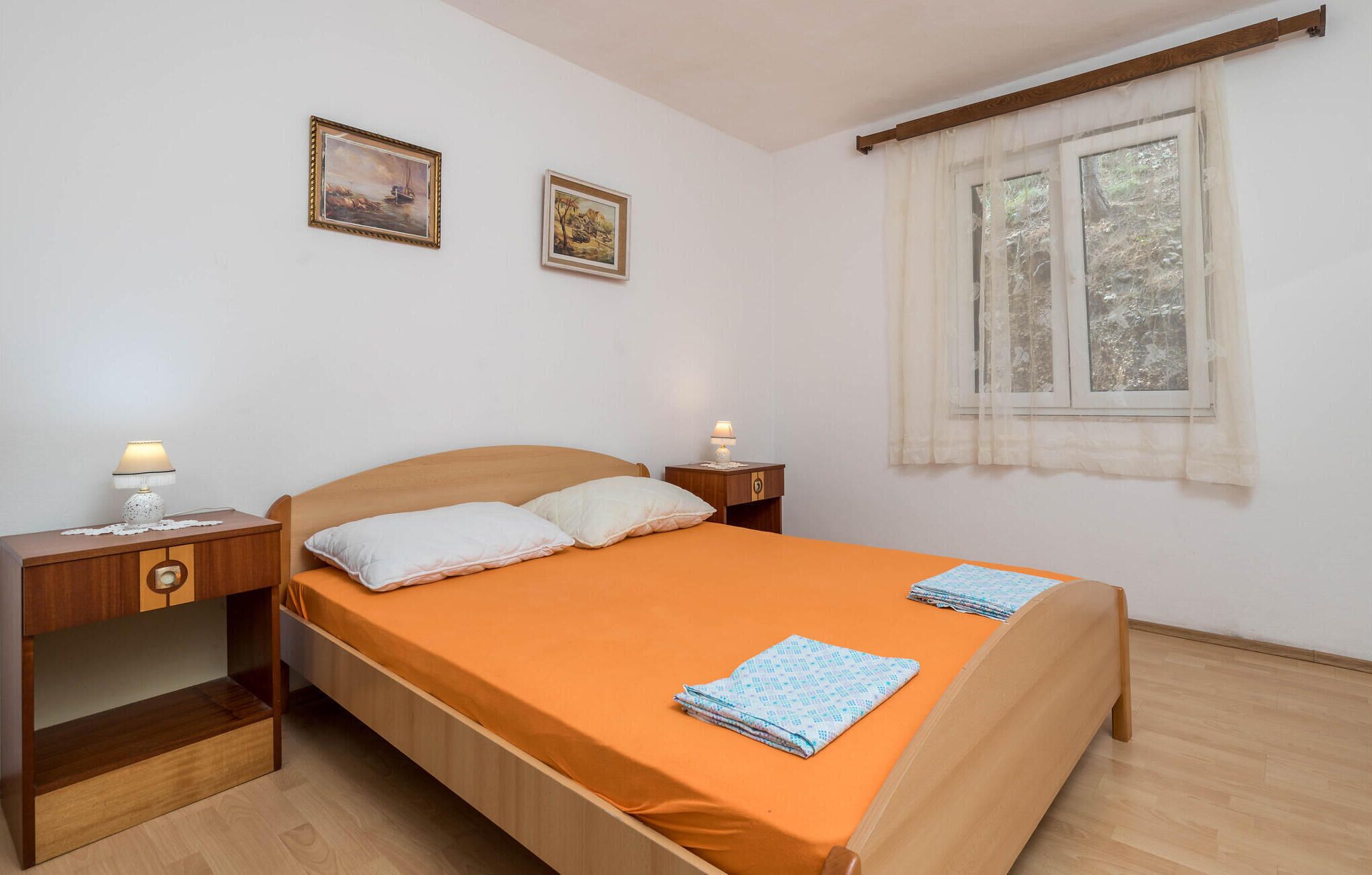 A bedroom with a wooden bed, two bedside tables with lamps, a window with sheer curtains, and two framed pictures on the wall.