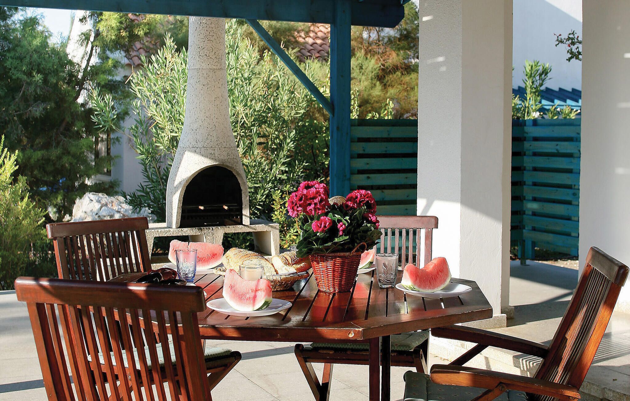 Een terras buiten met een stenen oven, houten stoelen, een tafel gedekt met brood, watermeloen en een mand vol bloemen.