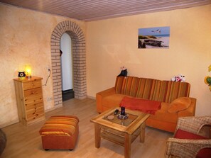 A cozy living room with a sofa, a wooden coffee table, a small ottoman, a wicker chair, and a lamp on a side table.