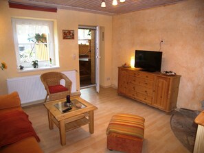 A cozy living room with a wicker chair, a wooden table, a television on a wooden cabinet, and a window with a view of a wooden deck.