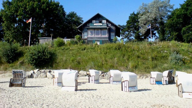 Een gebouw met twee verdiepingen en een balkon, omgeven door een grasheuvel en bomen. Strandstoelen staan op de voorgrond.