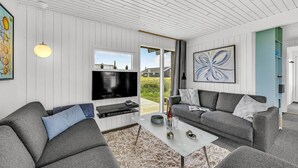 En moderne stue med et fladskærms-tv, grå sofaer, et sofabord og et stort vindue med udsigt til det fri.