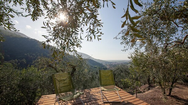 Two green chairs on a wooden deck overlooking a valley with olive trees and distant hills.