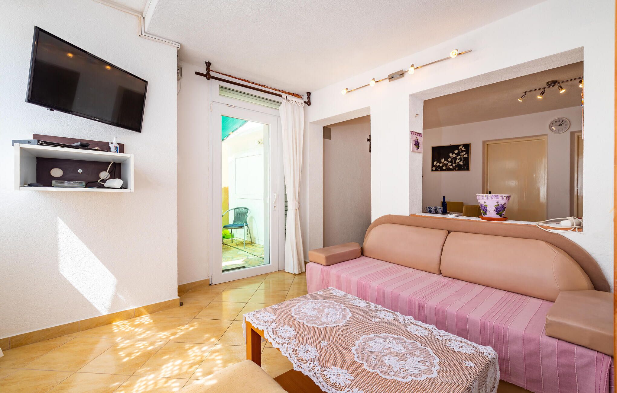 Un salon comprenant un canapé, un téléviseur fixé au mur, une petite table recouverte d’une nappe en dentelle et une porte donnant sur un espace extérieur.