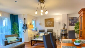 A living room with a sofa, a dining table, a lamp, a guitar, and a wall art piece. - Residence Bismarck App. Maris (Heringsdorf)