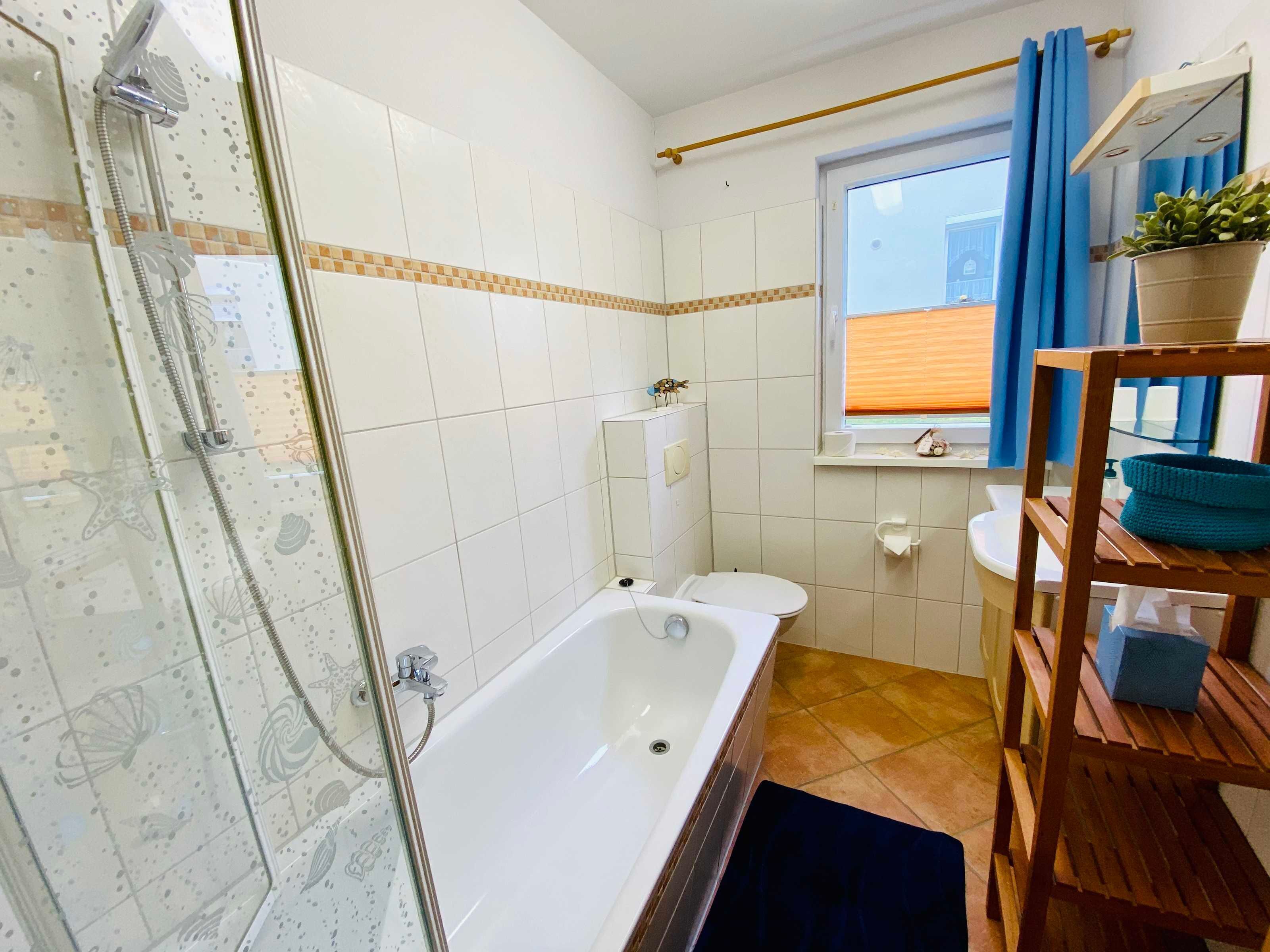 A bathroom with a glass-enclosed shower, a white bathtub, a toilet, and a window with blue curtains.