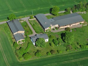 Une vue aérienne d’une ferme comprenant plusieurs bâtiments, dont une grande grange et des constructions plus petites, le tout entouré de champs verdoyants et d’arbres.