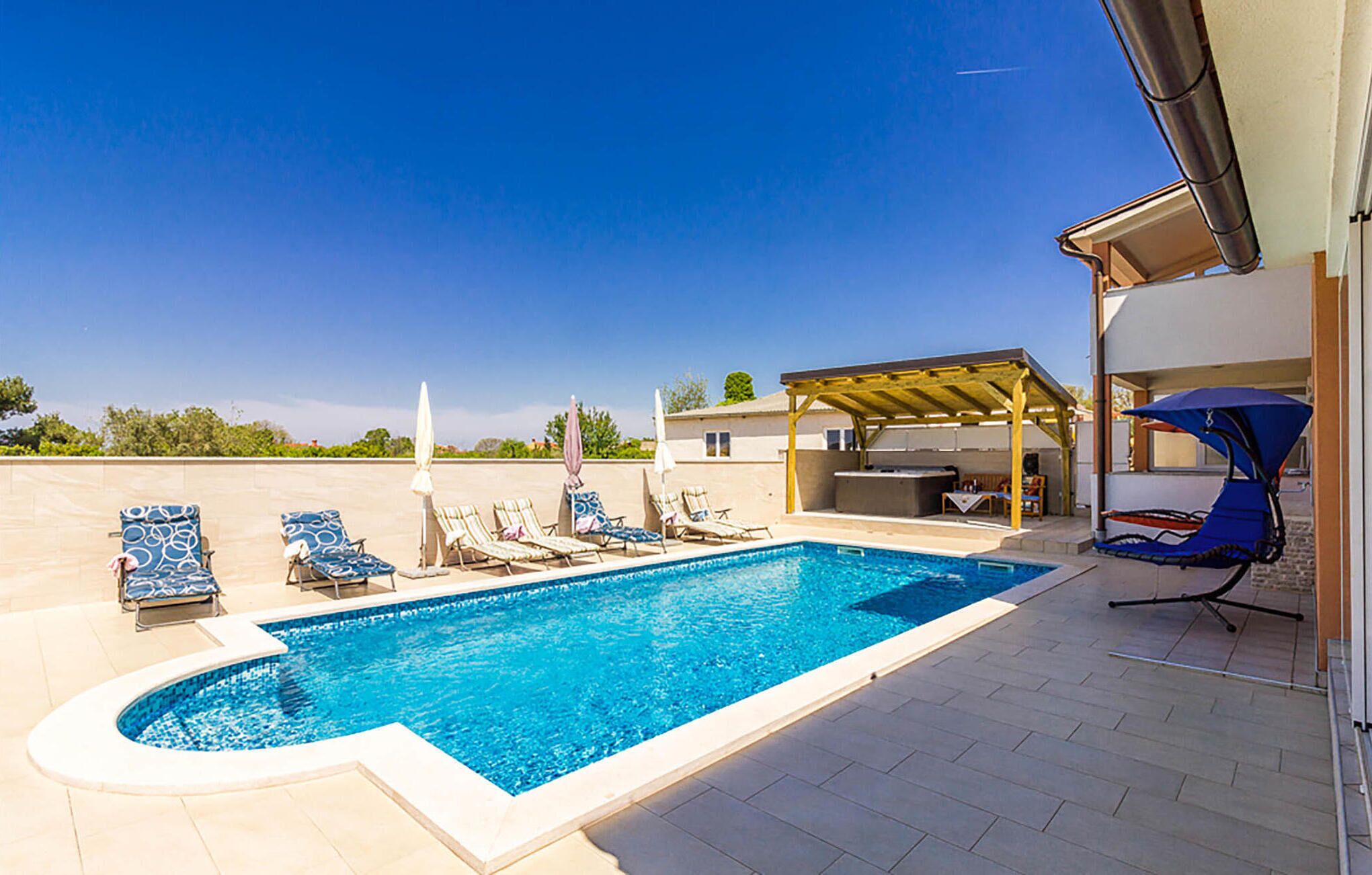 A swimming pool with lounge chairs and umbrellas, a covered patio area, and a view of the surrounding landscape.