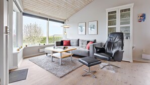 A modern living room with a grey sofa, a black leather chair, a wooden coffee table, and a large window offering a view of trees. - 3 bedroom nice home in Glesborg (Glesborg)