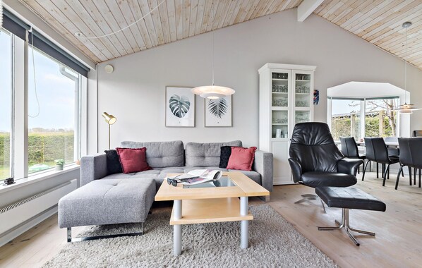 Una sala de estar moderna con un sofá seccional gris, una mesa de centro de madera y un sillón de cuero negro. Hay obras de arte con hojas enmarcadas en la pared, un aparador blanco con puertas de vidrio y un ventanal que ofrece vistas al exterior.