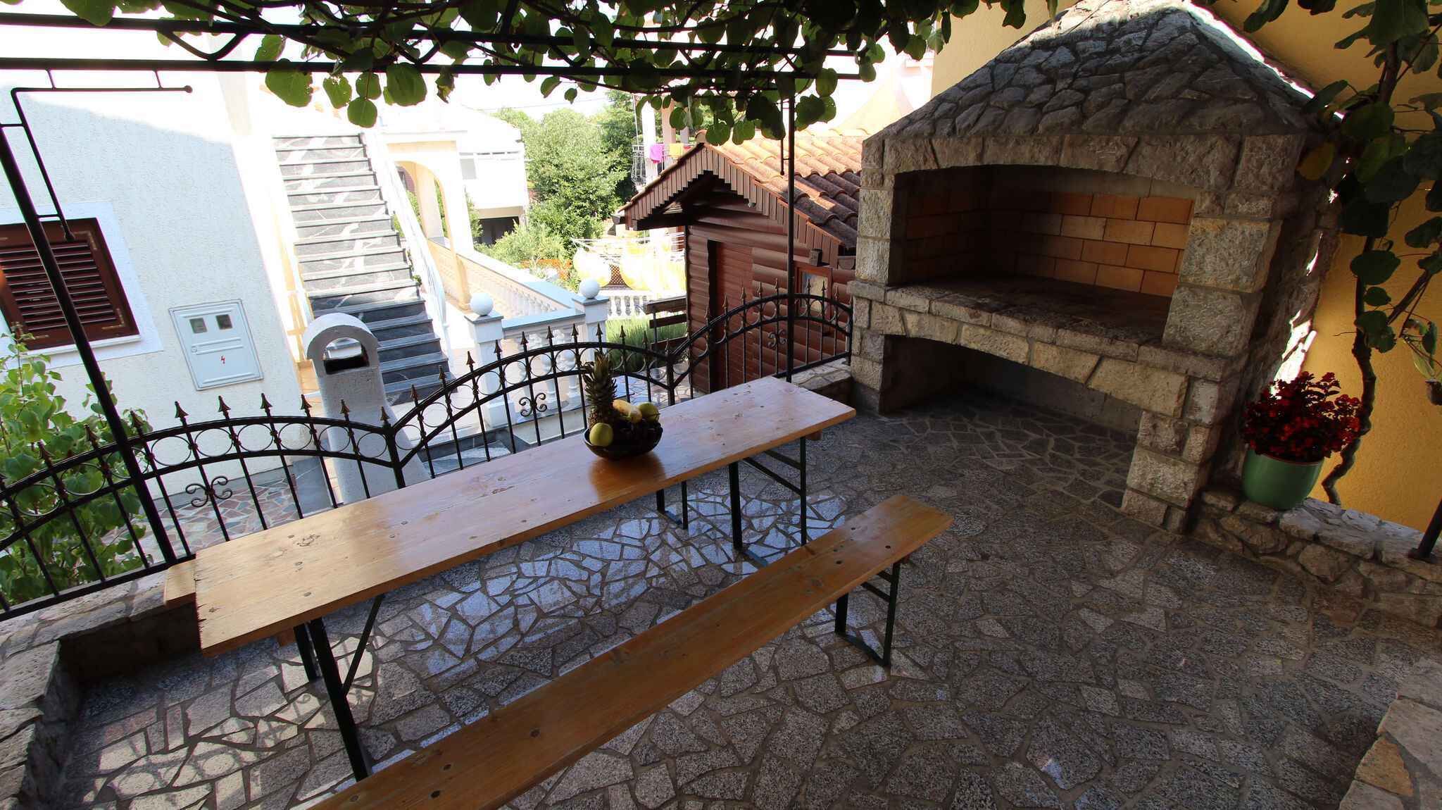 En stenbelagt terrasse med et træbord og bænke, en murstene ovn og et jerngitterhegn.