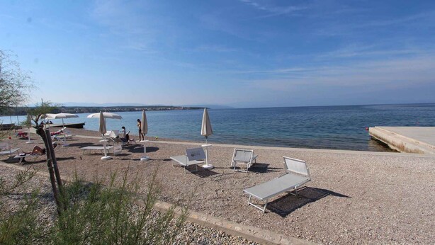 Ein Kiesstrand mit Liegestühlen und Sonnenschirmen, eine Betonpierde, die sich ins Wasser erstreckt, und freie Sicht auf das Meer.